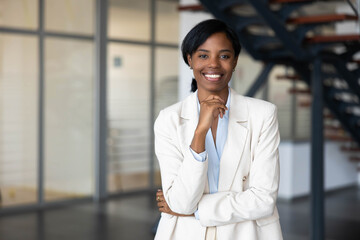 Obraz premium Black business woman standing confidently in a modern office environment, smiling at camera with hand on chin