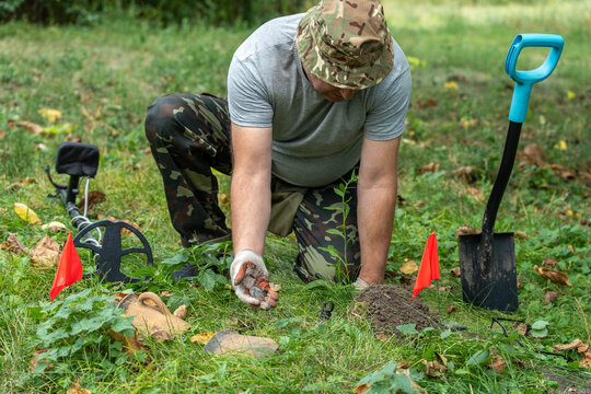 A person kneels on soft grass in a vibrant park, using a metal detector. Excitement fills the air as they uncover hidden items, marked by bright flags, surrounded by tools for digging
