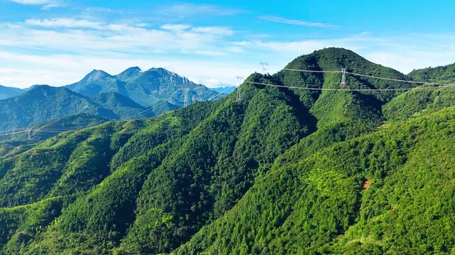 Lush mountain ridges covered in dense forest beneath a blue sky, with power lines crossing the slopes in bright sunlight and soft cloud cover.