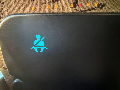 Seatbelt warning symbol displayed on a black seat in a vehicle, with patterned fabric visible in the background, indicating safety instructions for passengers