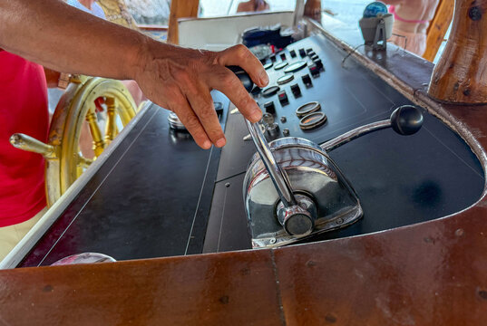Hand of a boat captain operating throttle control on a wooden boat dashboard with various instruments and steering wheel visible in the background