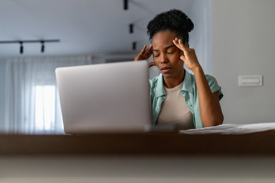 Black woman working at laptop in home office rubbing temples from stress and fatigue