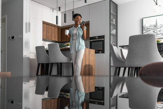 Morning routine for a black woman enjoying coffee while checking phone in modern kitchen