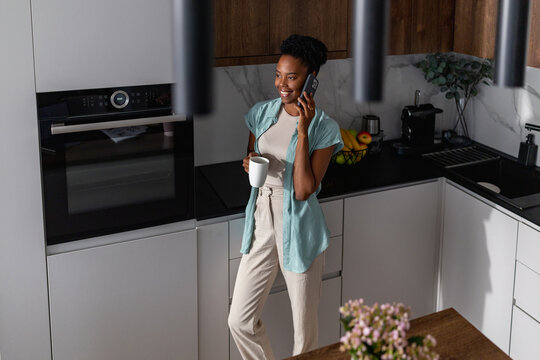 Woman talking on phone in modern kitchen holding a coffee mug and smiling