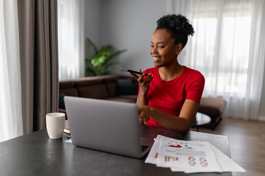 Smiling woman working from home on laptop while speaking into smartphone