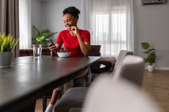 Young black woman smiling and checking phone while eating cherries at modern kitchen table