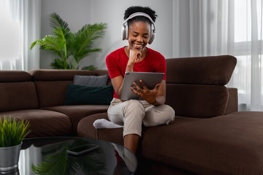 Young black woman relaxing on sofa with tablet and headphones, smiling while browsing at home