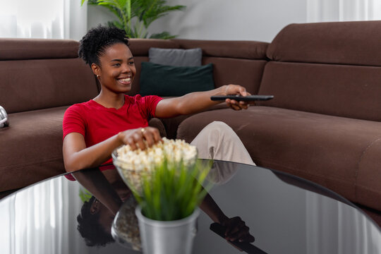 Relaxing on couch with popcorn and remote, black woman watches TV at home