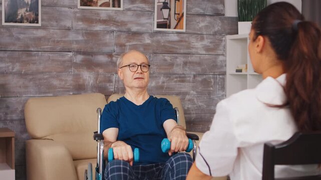 Older man in a wheelchair performs dumbbell arm exercises while a physiotherapist demonstrates movement during adaptive strength training and rehabilitation, while supporting mobility.