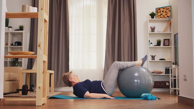 Senior woman exercises with a stability ball while lying on a yoga mat in a bright living room, practicing core strength, control and mobility at home, while supporting healthy aging.