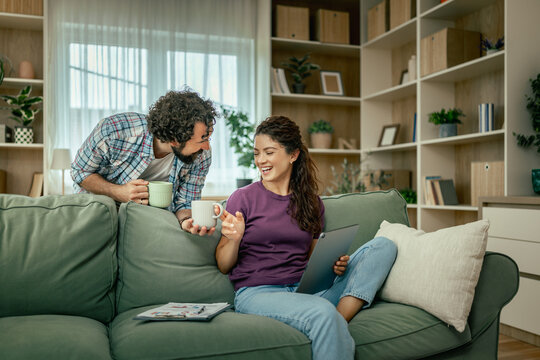Couple relaxing on sofa at home sharing coffee and laughing while using tablet