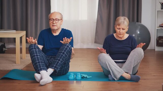 Older man and woman sit cross-legged with eyes closed and palms open, practicing controlled breathing and relaxation during a wellness routine at home, supporting mobility and healthy aging.