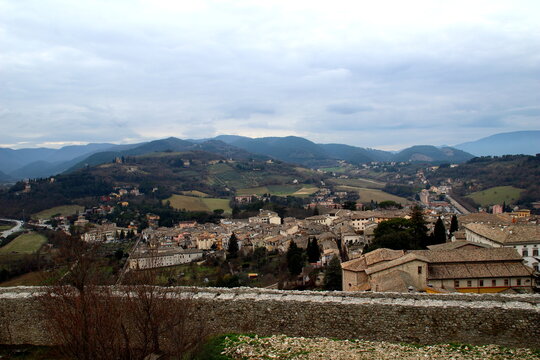 Moody view from a panoramic vantage point overlooking Spoleto, walls in the foreground, vintage buildings further down the hill, a vision of thickets, hills, roads, distant mountains, cloudy sky