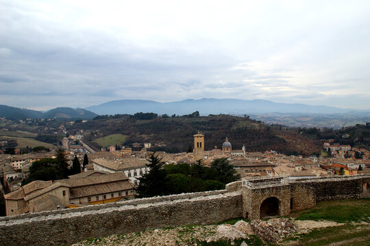 Mysterious view from the high stone ramparts of Rocca Albornoziana in Spoleto, traditional roofs of medieval houses and historic palazzos in the old town, rolling green hills and distant mountains