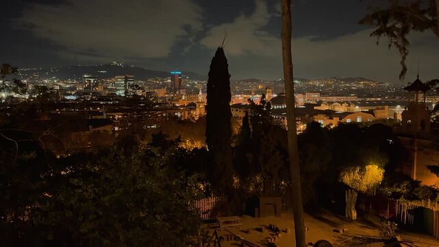 A stunning panoramic night view overlooking the illuminated urban city skyline of Barcelona, framed by lush trees and glowing streetlights under a dramatic, cloudy evening sky in Spain.

