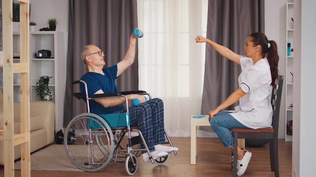Senior man in a wheelchair lifts dumbbells as a physiotherapist demonstrates upper-body movements during adaptive exercise, rehabilitation and active aging.