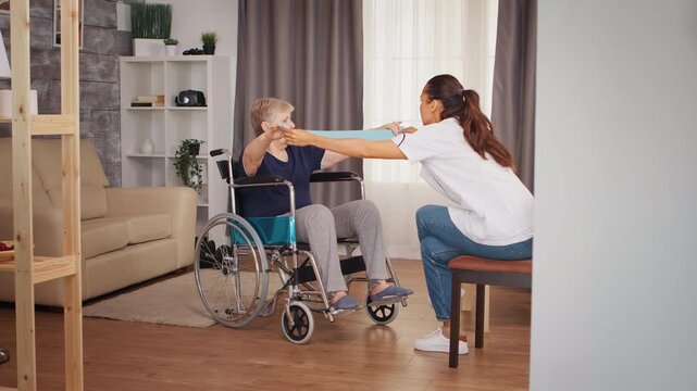Older wheelchair user reaches toward a resistance band held by a healthcare worker during guided rehabilitation, focusing on mobility, strength and assisted exercise, supporting strength training.
