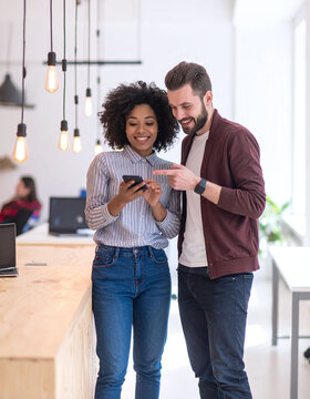 Diverse smiling colleagues look at a smartphone together in a bright modern office. A happy man and woman collaborate, sharing creative ideas in a warm tech startup environment.
