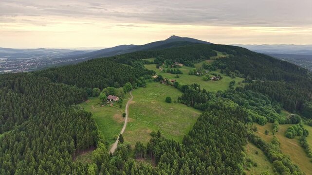 From high above, winding road links hilltop village over rolling green hills, surrounded by dense conifer forest and patches of fields and farms under soft pastel sky