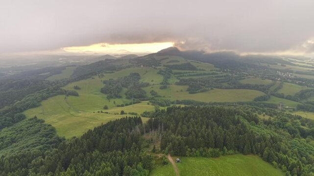 High aerial view captures patchwork fields across green rolling hills, dense woodland, and distant village settlement under dramatic cloud cover with sunbeams breaking through