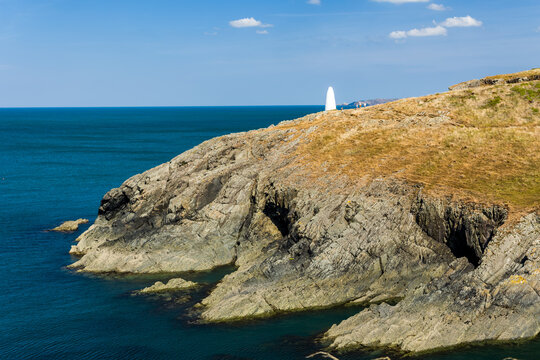 Elevated view of rocky headland, white navigation landmark on grassy ridge above craggy cliffs dropping into ocean, distant hikers