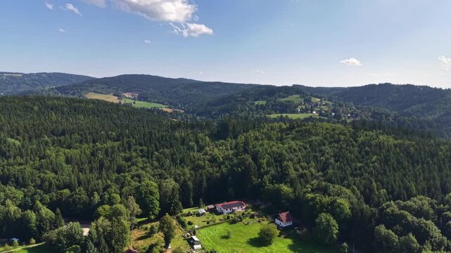 Wide aerial view shows rolling green hills covered with dense woods and fields, scattered village clusters, and red-roof houses stretching toward horizon beneath cloudless blue sky