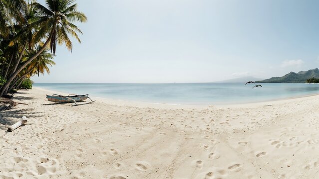 Panoramic view of tropical sandy beach with outrigger canoe on white shore, palm trees, calm ocean water, distant island mountain