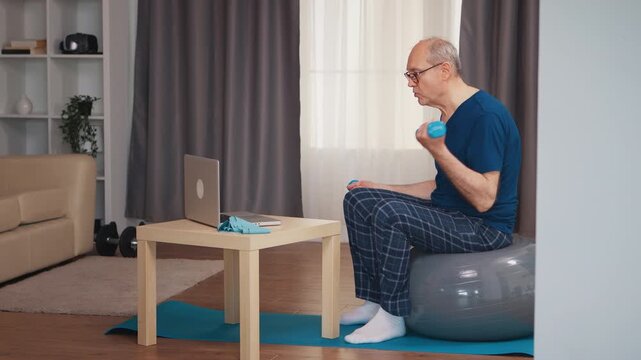 Senior man performs a seated dumbbell curl while watching an online workout on a laptop, showing focused strength training and active aging at home.
