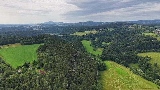 Gentle wide aerial view shows vibrant patchwork farmland, rolling grassy hills, and forest ribbons leading toward small village on horizon under bright sunlight and soft cloud cover