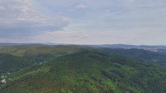 High aerial view over lush green hills and forest canopy, rolling valleys and ridges stretching toward distant city edge under blue sky and gentle clouds, calm scenic landscape