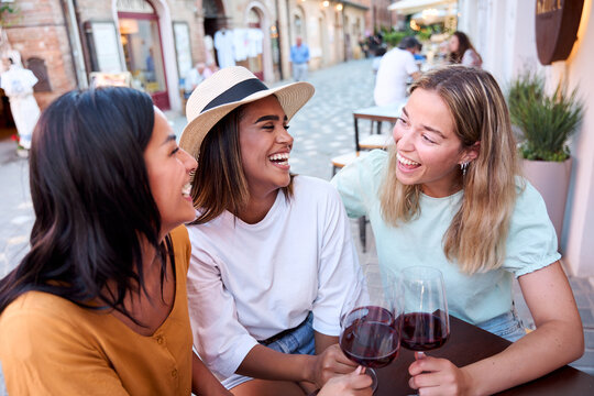 Three cheerful multi-ethnic female friends are happily toasting with glasses of red wine while enjoying their vacation together in an outdoor setting in an italian city