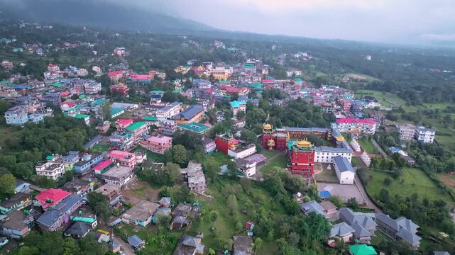 Wide aerial flyover above the vibrant town of Bir, Himachal Pradesh, highlighting traditional monastery rooftops and misty mountain backdrops