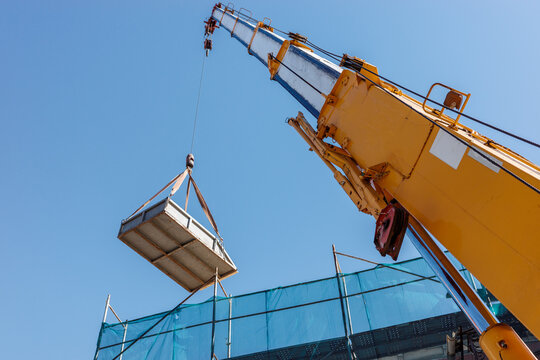 Construction crane lifts steel platform above building site, heavy machinery handling suspended load during industrial installation, engineering and development.