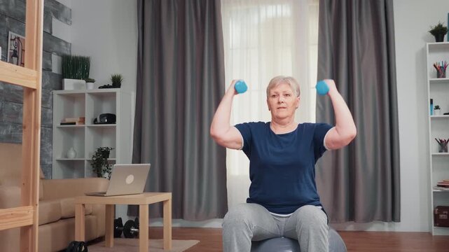 Senior woman performs seated dumbbell shoulder exercises on a stability ball at home, supporting upper-body strength, balance and functional fitness for active aging, emphasizing mobility.