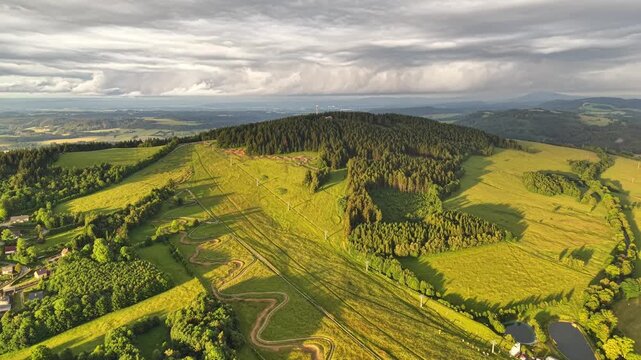 Drone capture reveals forest crowned hill with tall summit tower, bordered by lush pastures and winding paths, village edges visible at bottom left, while heavy clouds cast soft light and long