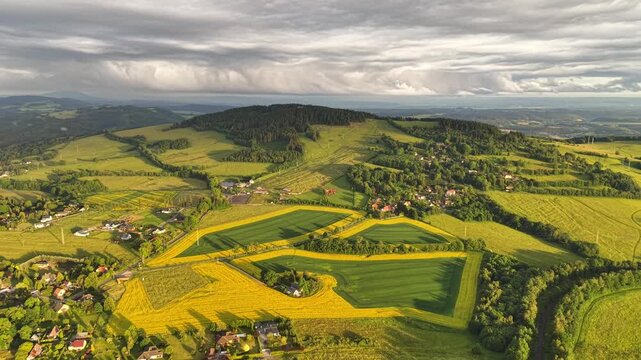 Panoramic aerial scene of patchwork farmland in vivid green and yellow tones, showing hedgerows, tree lines, winding roads, and clustered village settlements across open rural terrain