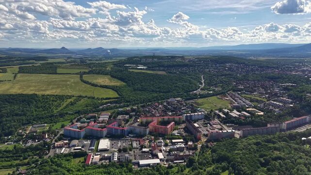 high altitude view over valley town featuring colorful housing blocks, patchwork farmland, and surrounding forests, with visible roads, rooftops, and distant hills beneath clear blue sky