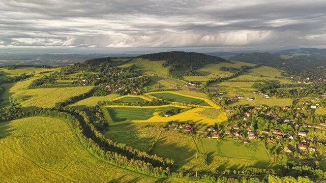 Sunlit countryside landscape from above, showing patchwork fields, hedgerows, winding roads, and forested ridges with scattered village clusters under dramatic layered clouds