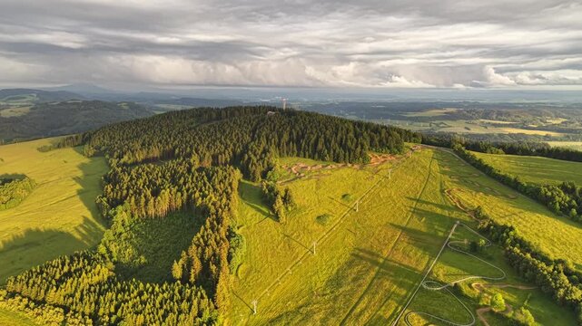 Wide aerial landscape shows forest ridge surrounded by pasture and meadow, green fields with winding paths, distant farmland and rolling valley, lit by warm golden hour sunlight and cloud shadow
