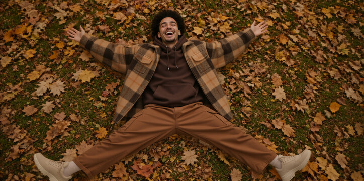 Happy young man lying on autumn leaves. Cheerful mixed race male laughing with arms outstretched in park. Carefree fall lifestyle concept