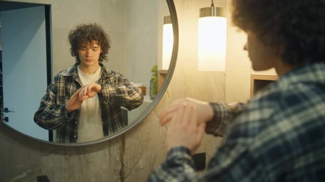 A curly-haired man in a checkered shirt stands in a modern bathroom, applying cream to his hands while facing a round mirror.