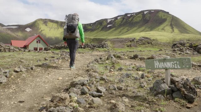 A lone hiker treks along the famous Laugavegur trail in Iceland, passing the Hvannagil camp site. Remote volcanic landscape with green mountains and snow patches under an overcast sky.