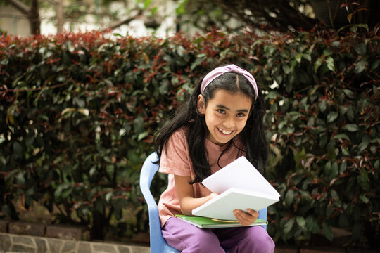 Young girl reading a book outdoors while sitting in a chair, enjoying a quiet moment of childhood learning, imagination and independent reading.