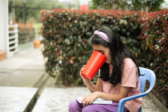 Young girl taking a drink from a red cup while reading outdoors, combining childhood leisure, hydration and independent reading in a relaxed setting.