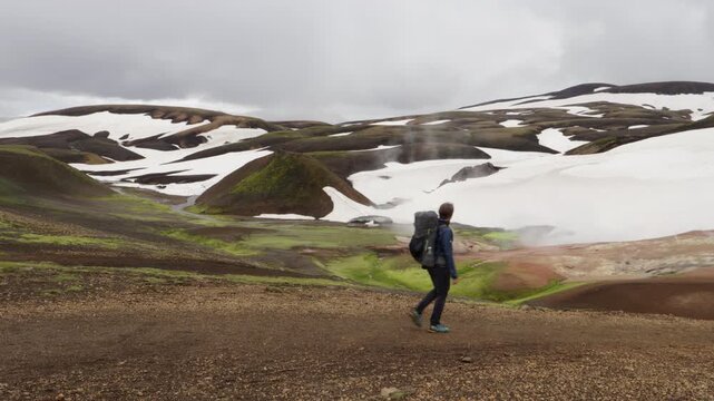 A lone hiker with a backpack traverses the Laugavegur trail on Iceland, a rugged volcanic landscape dotted with geothermal steam and snowfields under a cloudy sky.