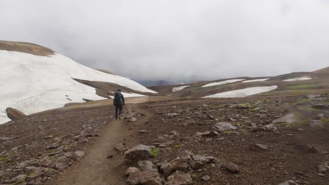A lone hiker treks along the famous Laugavegur trail on Iceland, navigating a vast and rugged volcanic landscape under a cloudy sky. Majestic mountains and snow patches add to the remote scenery.