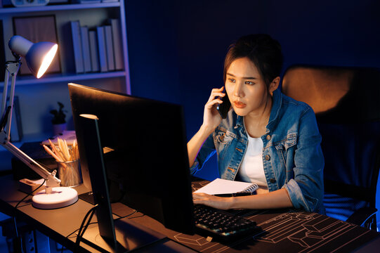 Young beautiful Asian creative woman calling on phone desk to customer or coworker explaining creator project's high profit while searching market analysis on pc at neon office at night. Stratagem.
