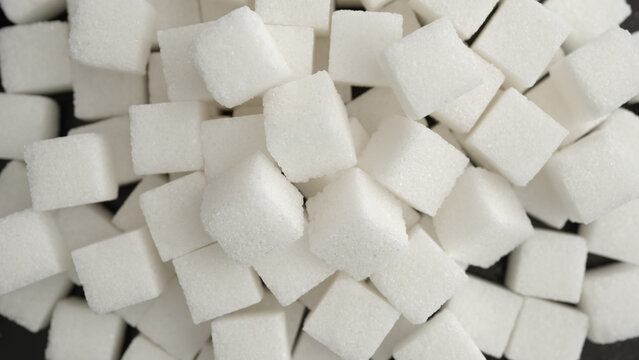 White sugar cubes piled together in an overhead close-up, scattered across a dark surface with visible crystalline texture.
