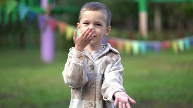 Cute smiling boy in plaid jacket performs the viral six seven meme hand gesture outdoors on grass at a kids party with colorful flags.