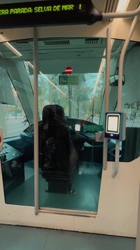 Interior view of a driver operating a modern electric city tram through urban streets, filmed from behind the transparent glass cabin partition inside the passenger compartment during daytime.

